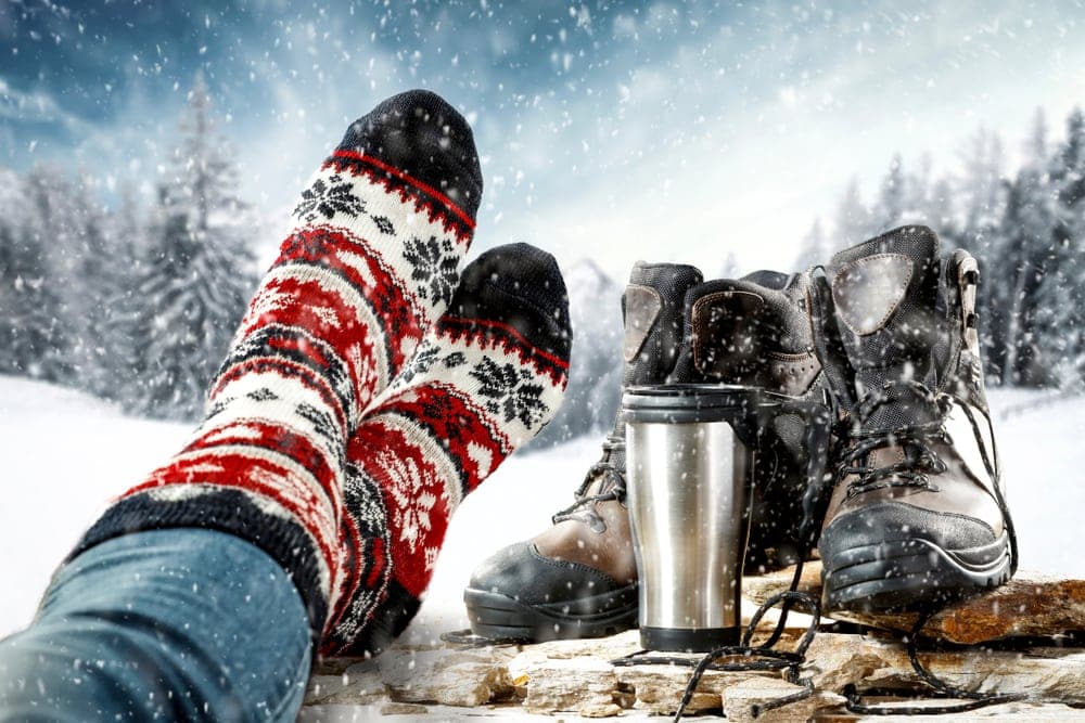 Person wearing a christmas-designed heated socks with boots and tumblr besides during winter camping