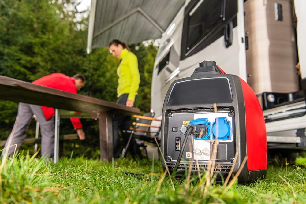 A camping generator with a background of an RV and a couple setting up a campsite