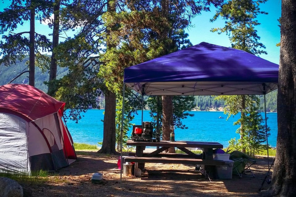 Red and white dome tent beside a picnic table with stove and lantern under a purple canopy, with a blue lake beyond pines